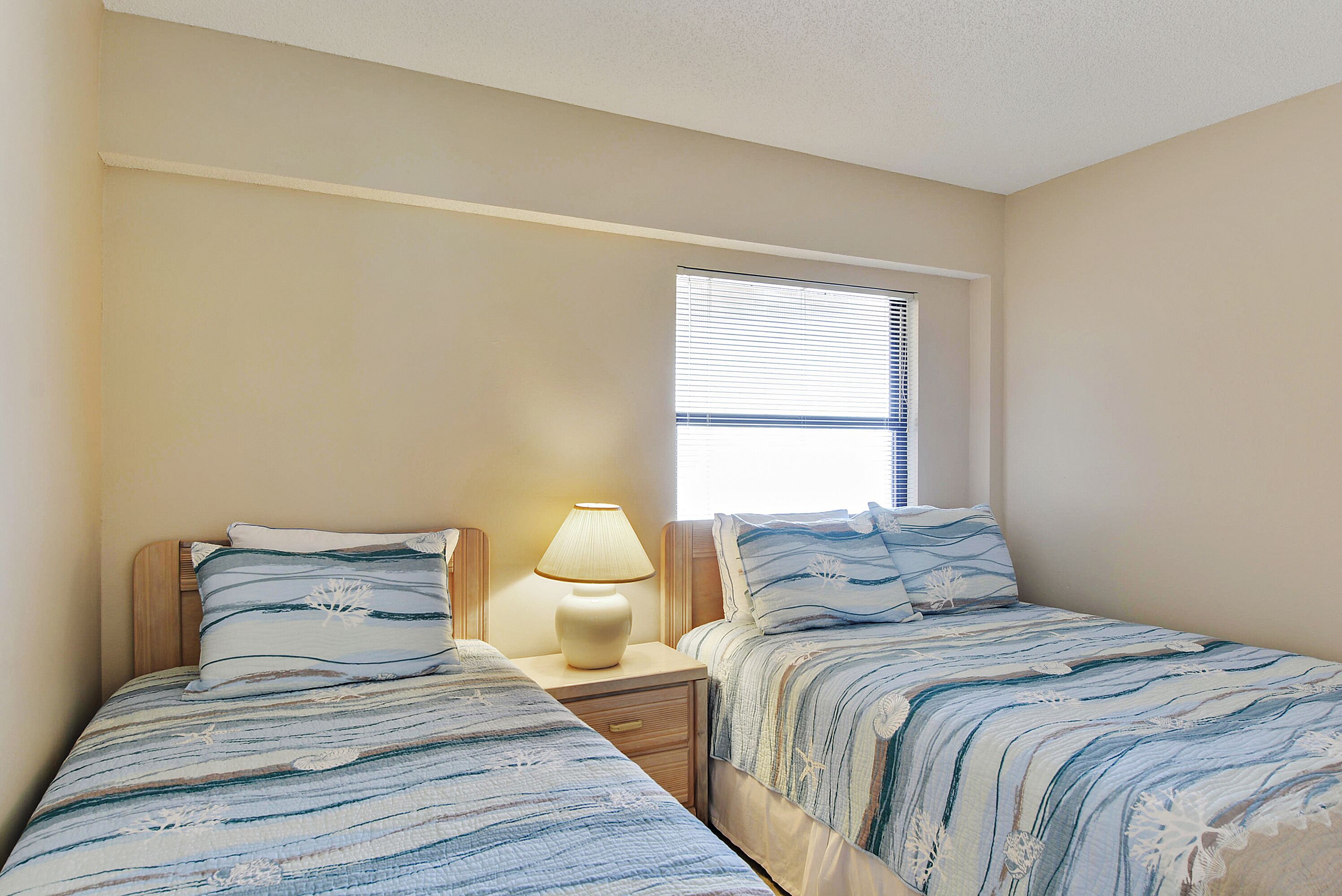 Une chambre avec deux lits simples, une table de chevet avec une lampe et une fenêtre avec des stores.