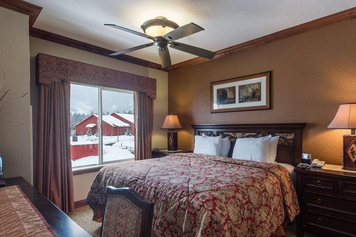A hotel room with a large bed, a ceiling fan, a window with curtains, a framed picture on the wall, and a nightstand with a lamp.