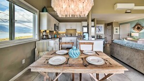 Un comedor con una mesa de madera dispuesta para cuatro personas, un jarrón azul como centro de mesa y una cocina al fondo con armarios blancos y una lámpara colgante.