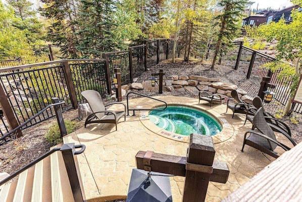 A hot tub surrounded by stone seating and a wooden deck, with a metal fence and trees in the background.