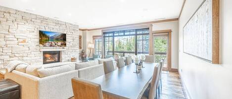 A spacious living room with a stone fireplace, a large TV mounted on the wall, and a dining area with a long table and chairs.