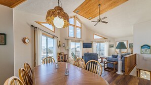 Un comedor amplio con una gran mesa y sillas de madera, un ventilador de techo y ventanas grandes que ofrecen vistas al exterior.