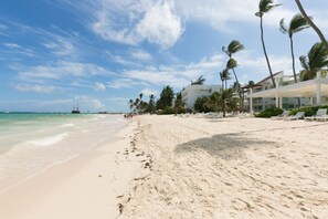 A beach with white sand, palm trees, and a building with a white roof and balconies.