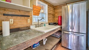 Una cocina con un refrigerador de acero inoxidable, paredes de madera, un lavamanos blanco y una ventana con persianas.