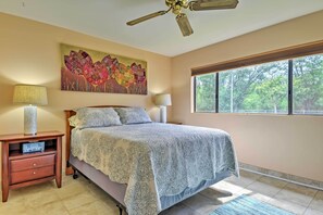 A bedroom with a bed, nightstands, lamps, a ceiling fan, a large window, and a painting on the wall. - Kihei Condo: Steps to Kalepolepo Beach Park! (Kihei)