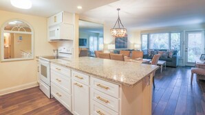 A spacious kitchen with white cabinets and a granite countertop, a dining area with a sofa and chairs, and a large window providing natural light.