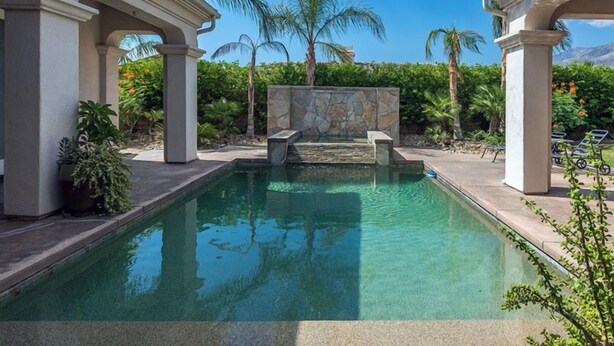 Une piscine rectangulaire avec des marches en pierre menant à l’intérieur, entourée de palmiers et d’un mur de pierre.