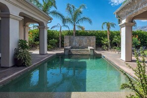 Una piscina rectangular con escalones de piedra para entrar, rodeada de palmeras y un muro de piedra.