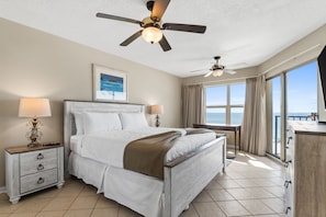 A hotel room with a bed, two bedside tables, a ceiling fan, a window with curtains, and a view of the ocean.