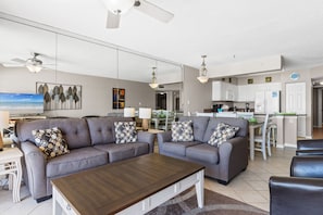 Una sala moderna con dos sofás, una mesa de centro y un televisor. Se ve una cocina al fondo, con armarios blancos y un comedor.