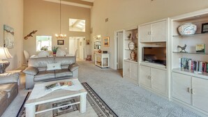 Amplia sala de estar con techo alto, un sofá grande, una mesa de centro y un televisor. Se observa una estantería con diversos objetos y una puerta que conduce a otra habitación.