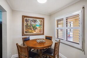 Un coin repas avec une table ronde en bois, quatre chaises, un tableau encadré représentant une scène de plage tropicale et une fenêtre avec des stores.