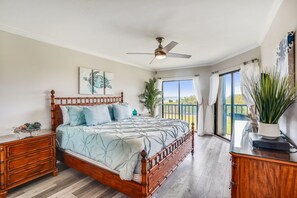 Une chambre avec un lit en bois, un ventilateur de plafond, une grande fenêtre avec des rideaux et une vue sur l’extérieur.