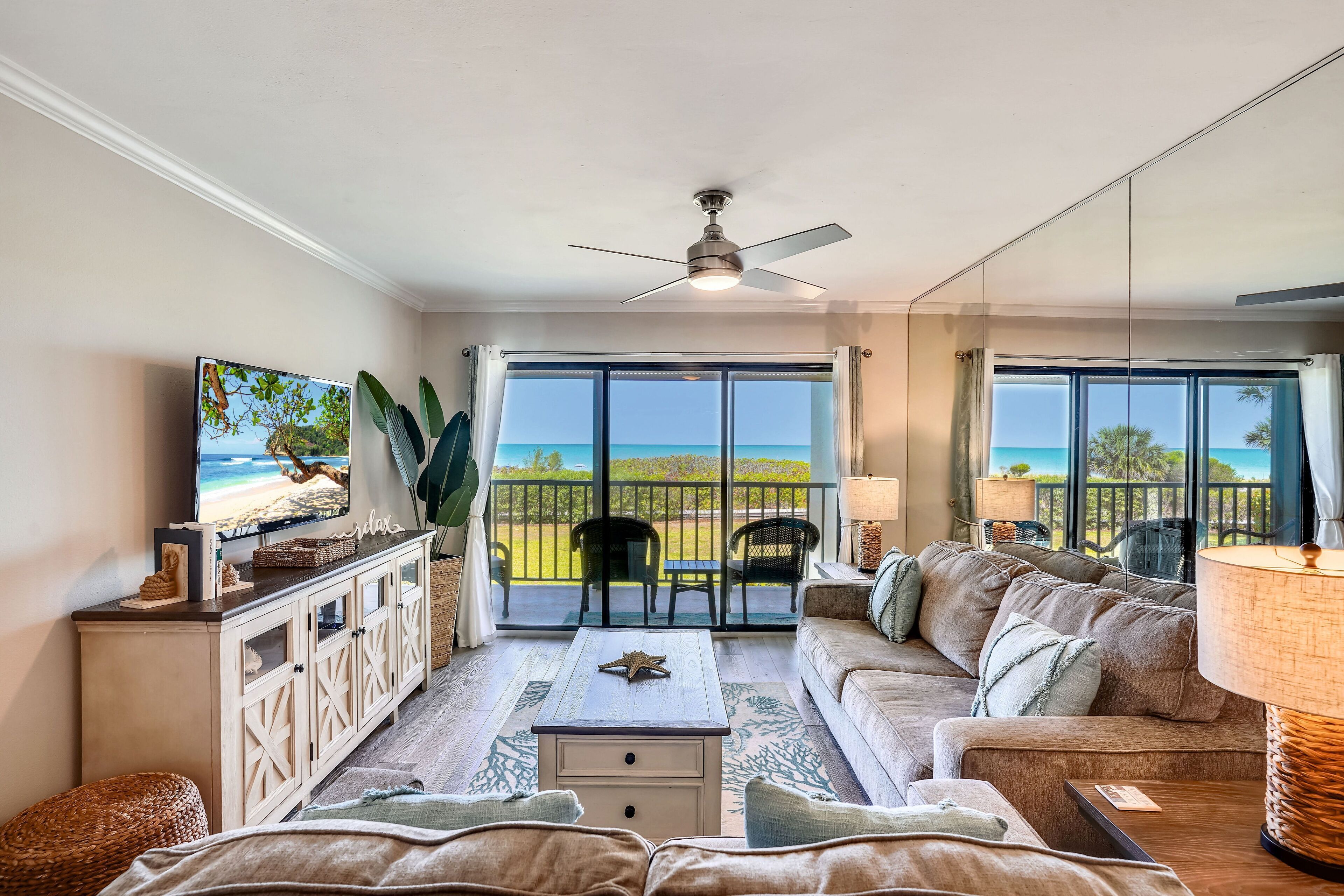 A living room with a sofa, coffee table, and TV. There is a balcony with chairs and a view of the ocean.