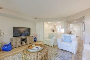 Una sala de estar con televisor, sofás blancos, una mesa de centro y un otoman de mimbre.