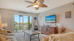 Ein Wohnzimmer mit Deckenventilator, Fernseher, Esstisch und Meerblick durch große Fenster.