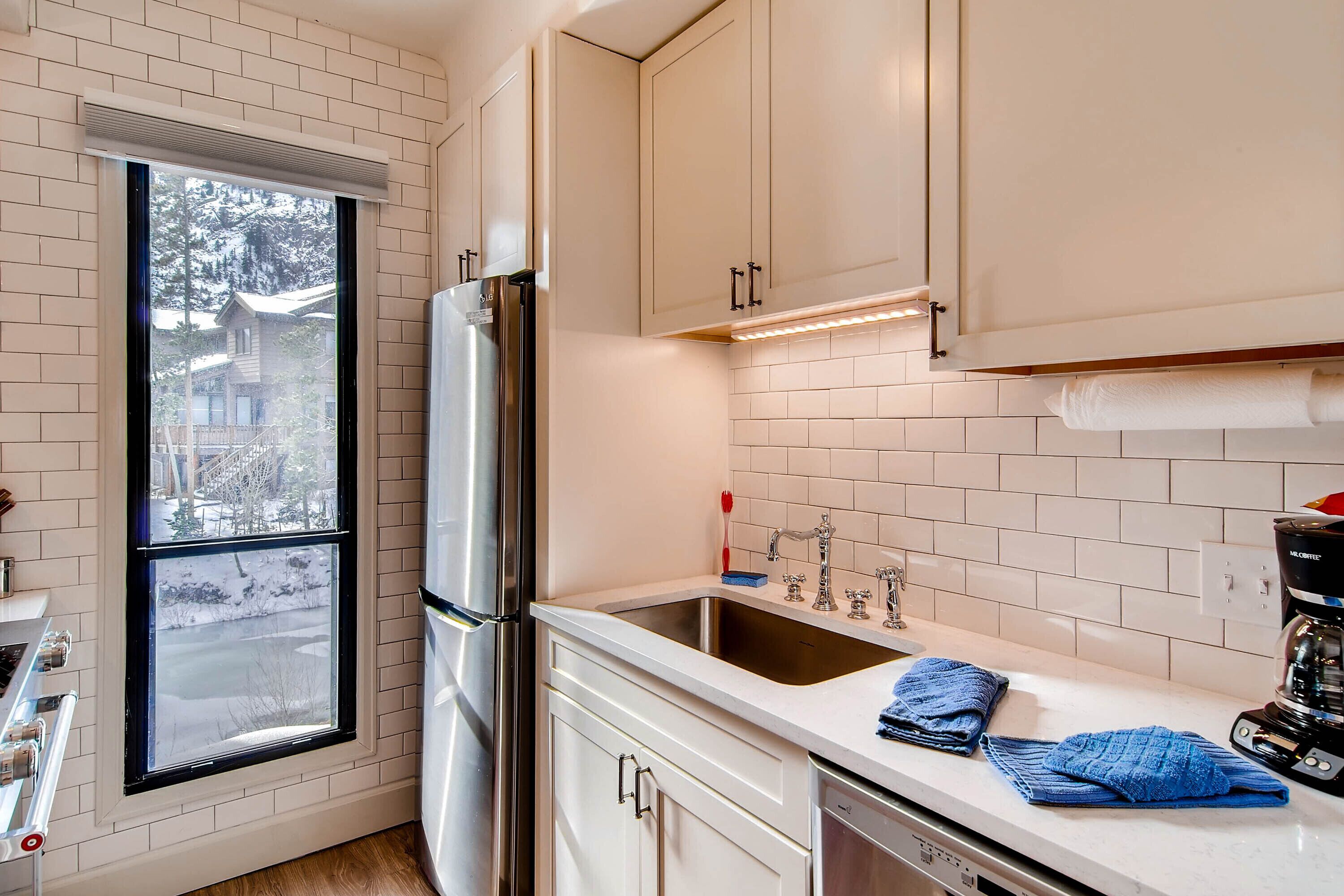 A kitchen with white cabinets, a stainless steel refrigerator, a sink, and a window with a view of a snowy landscape.