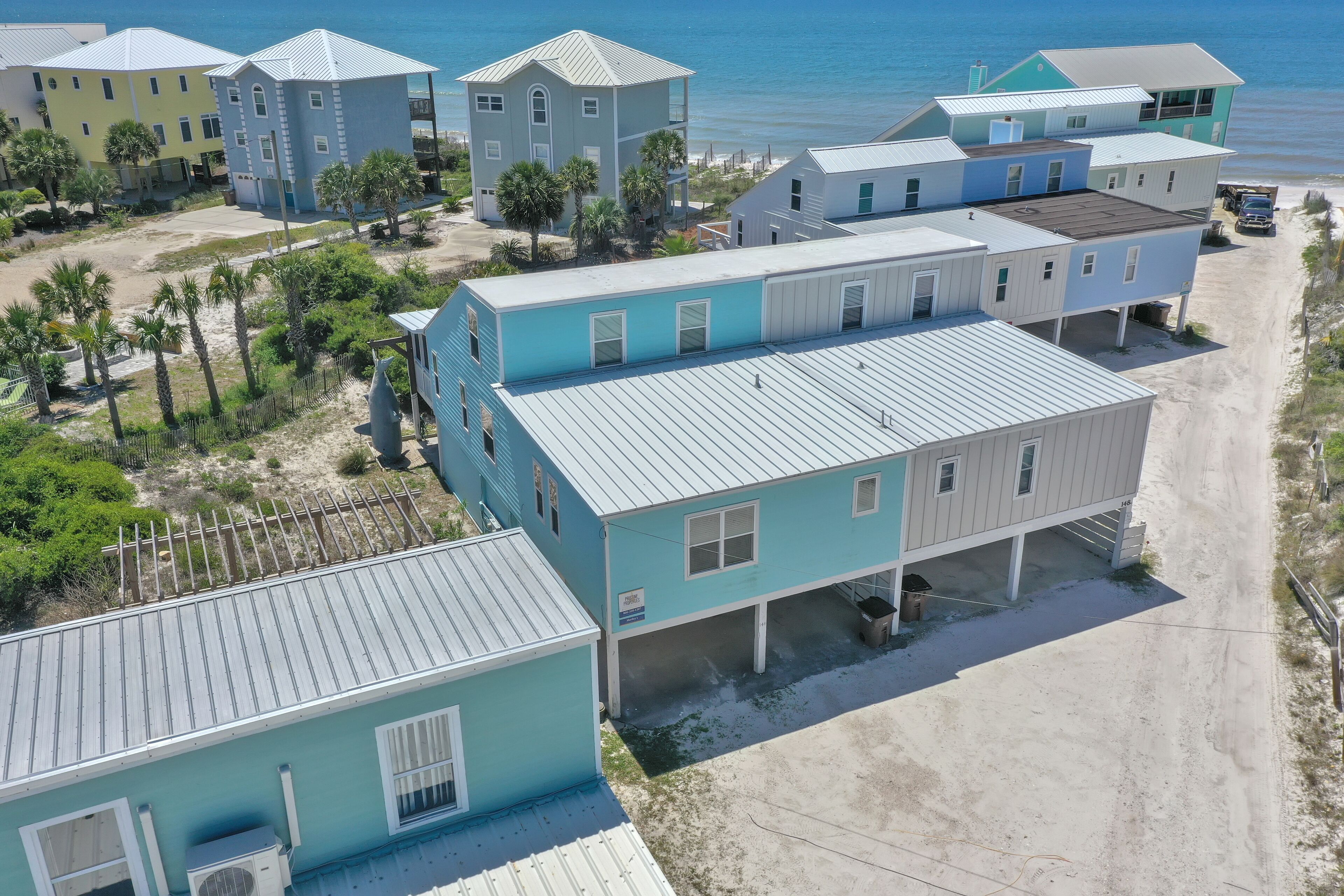 A coastal residential area with multiple two-story houses, featuring a mix of light blue and yellow exteriors, surrounded by palm trees and a sandy beach.
