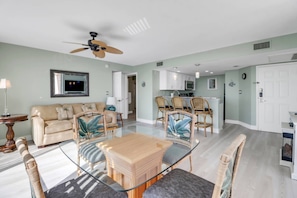 Un vaste espace de vie comprenant un canapé, une table basse en verre, des fauteuils en osier, un ventilateur de plafond et, au fond, une cuisine.