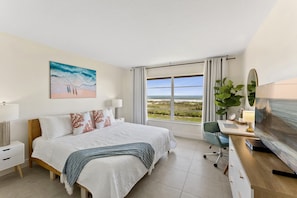 A hotel room with a large bed, a desk with a computer, a TV, a window with a view of the beach, and a painting on the wall.