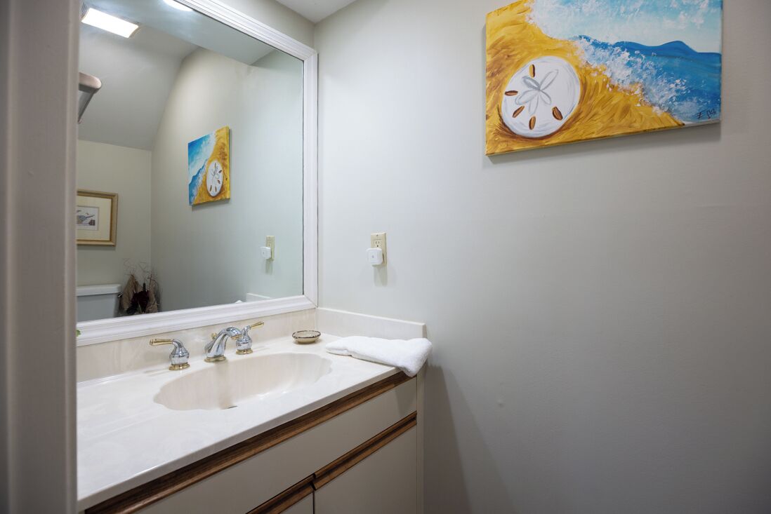 Une salle de bain avec un lavabo blanc, deux robinets et un miroir. Il y a un tableau au mur et une photographie encadrée au-dessus du miroir.