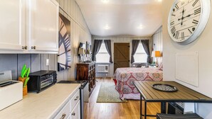 A compact room with a kitchenette, a bed with a patterned bedspread, and a large wall clock. - Historic 'Hardy House' on Main Street w/ Fire Pit! (Hardy)