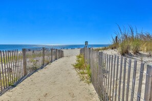 Un sentier de sable bordé d’une clôture en bois d’un côté et d’une clôture en métal de l’autre, qui mène à l’océan.