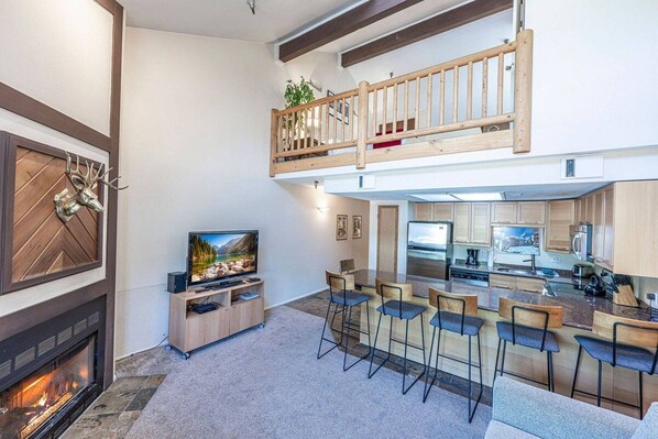 A modern interior with a fireplace, a television, a kitchen area, and a dining space with bar stools.