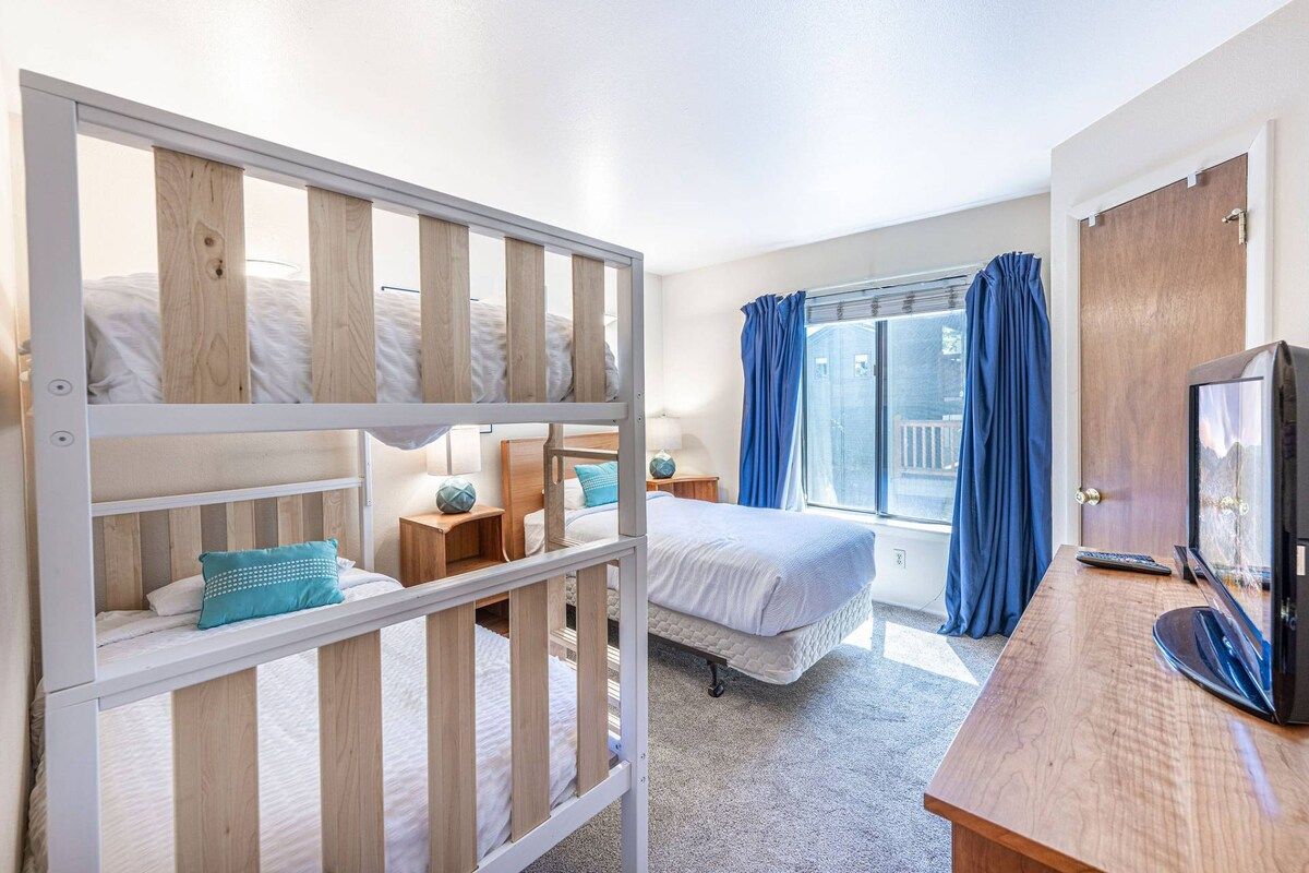 A bedroom with a bunk bed, a single bed, a desk with a television, and a window with curtains.