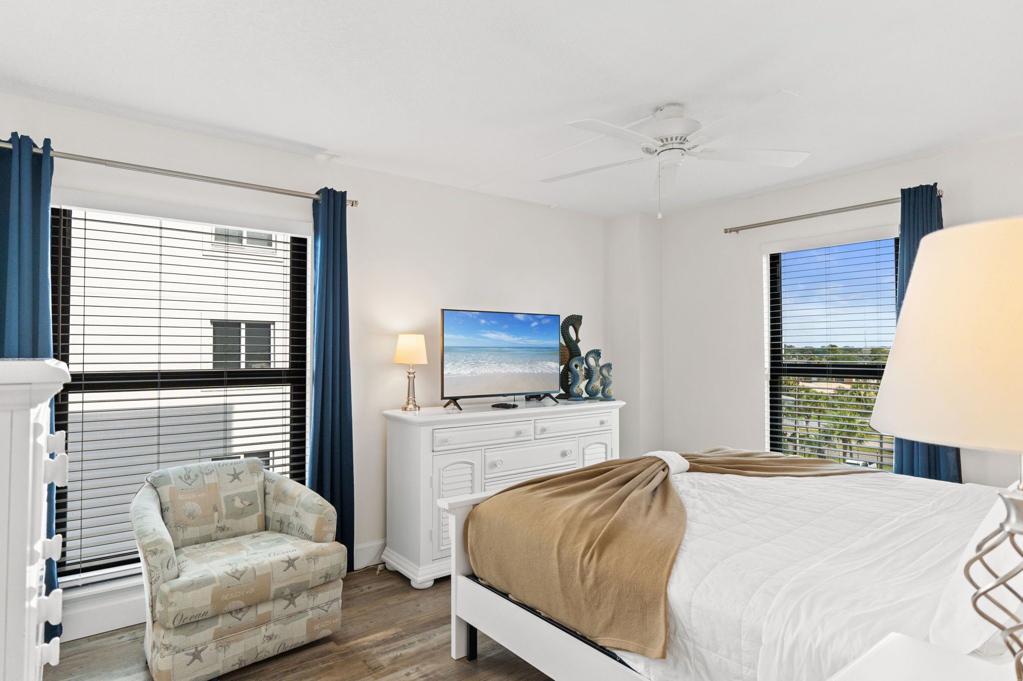 A hotel room with a bed, a TV, a dresser, a chair, and large windows with blinds.