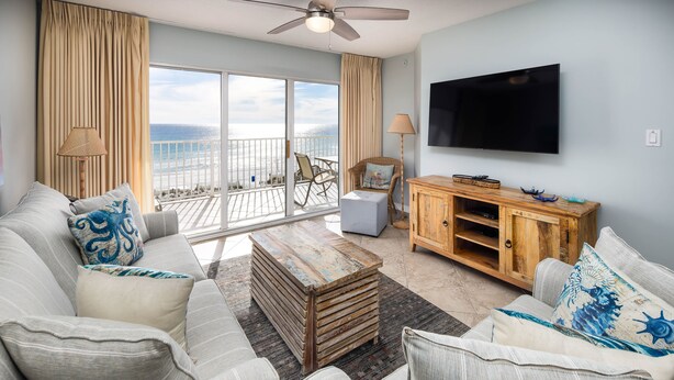 A spacious living room with a large window overlooking the ocean, a ceiling fan, a flat-screen TV, and a wooden cabinet.