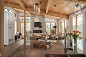 A spacious kitchen with a central island, wooden ceiling beams, large windows, and a fireplace.