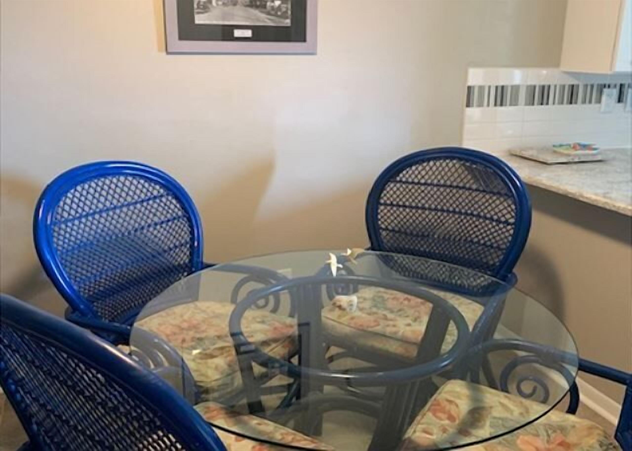 Una mesa redonda de vidrio con cuatro sillas azules, cada una con un cojín floral. Encima de la mesa, hay un cuadro enmarcado colgado en la pared.