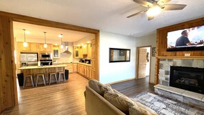 A modern kitchen with wooden cabinets, a central island, and bar stools. There is a fireplace with a stone surround and a television mounted above it. - Greenway Crossing @ Spring Brook Resort (Wisconsin Dells)