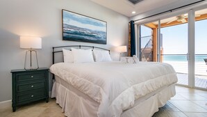 Un dormitorio con una cama grande, una mesita de noche, un ventilador de techo y vista a la playa a través de una puerta abierta.