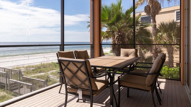 Un patio iluminado por el sol con una mesa y sillas de madera, con vista a una playa y palmeras.
