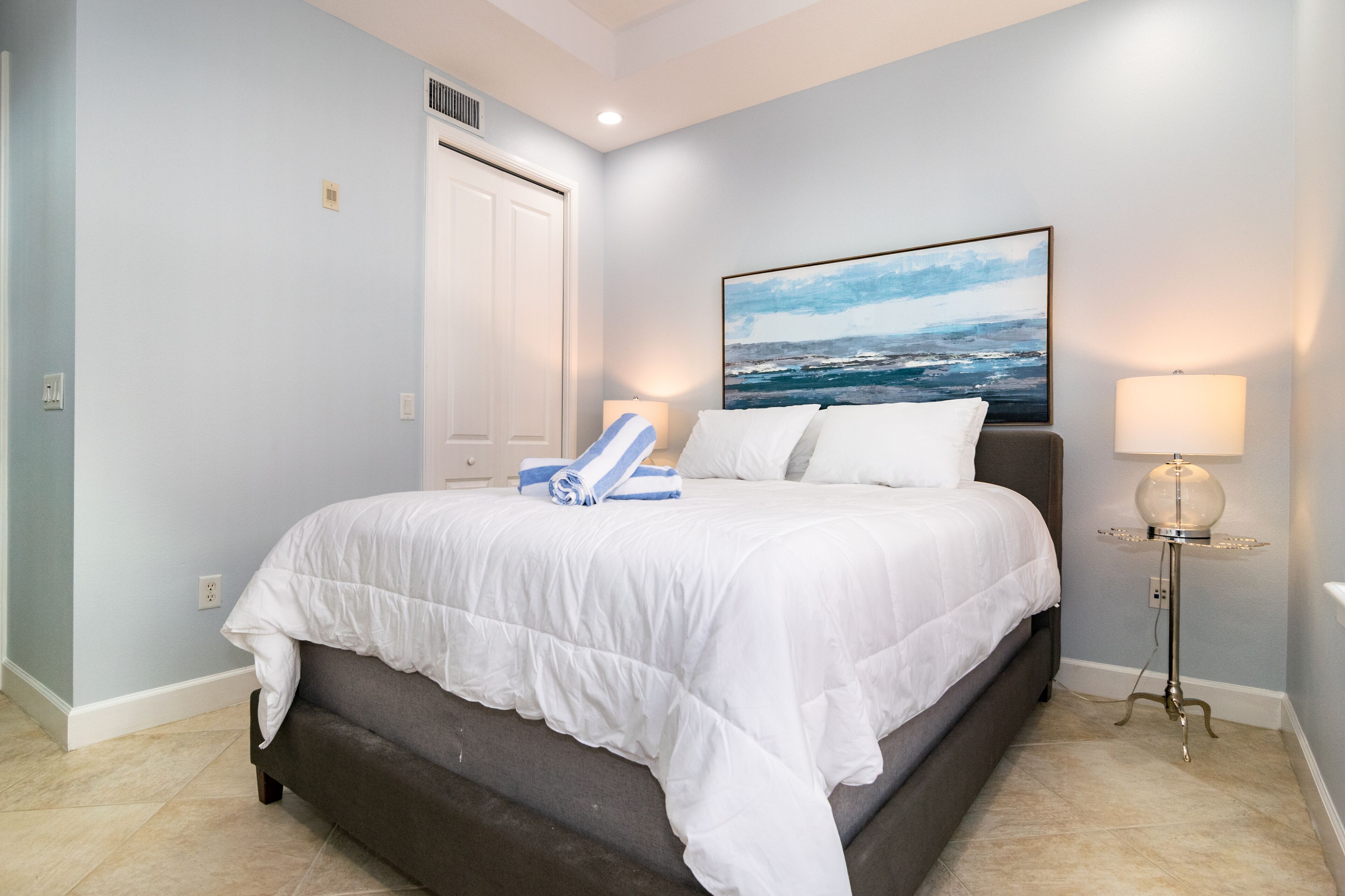 A bedroom with a bed, a painting of the ocean, a lamp, and a door.