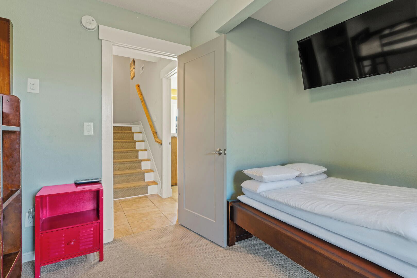 Une chambre avec un lit, un téléviseur fixé au mur, un meuble de rangement rouge et un escalier menant à une autre pièce.