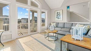 A spacious living room with a large sliding glass door leading to a balcony, a grey sofa, a wooden coffee table, and a framed artwork on the wall. - Coastal Calling at Corolla Shores 7 Bedroom Home, Near Beach Access + Private Pool (Corolla)