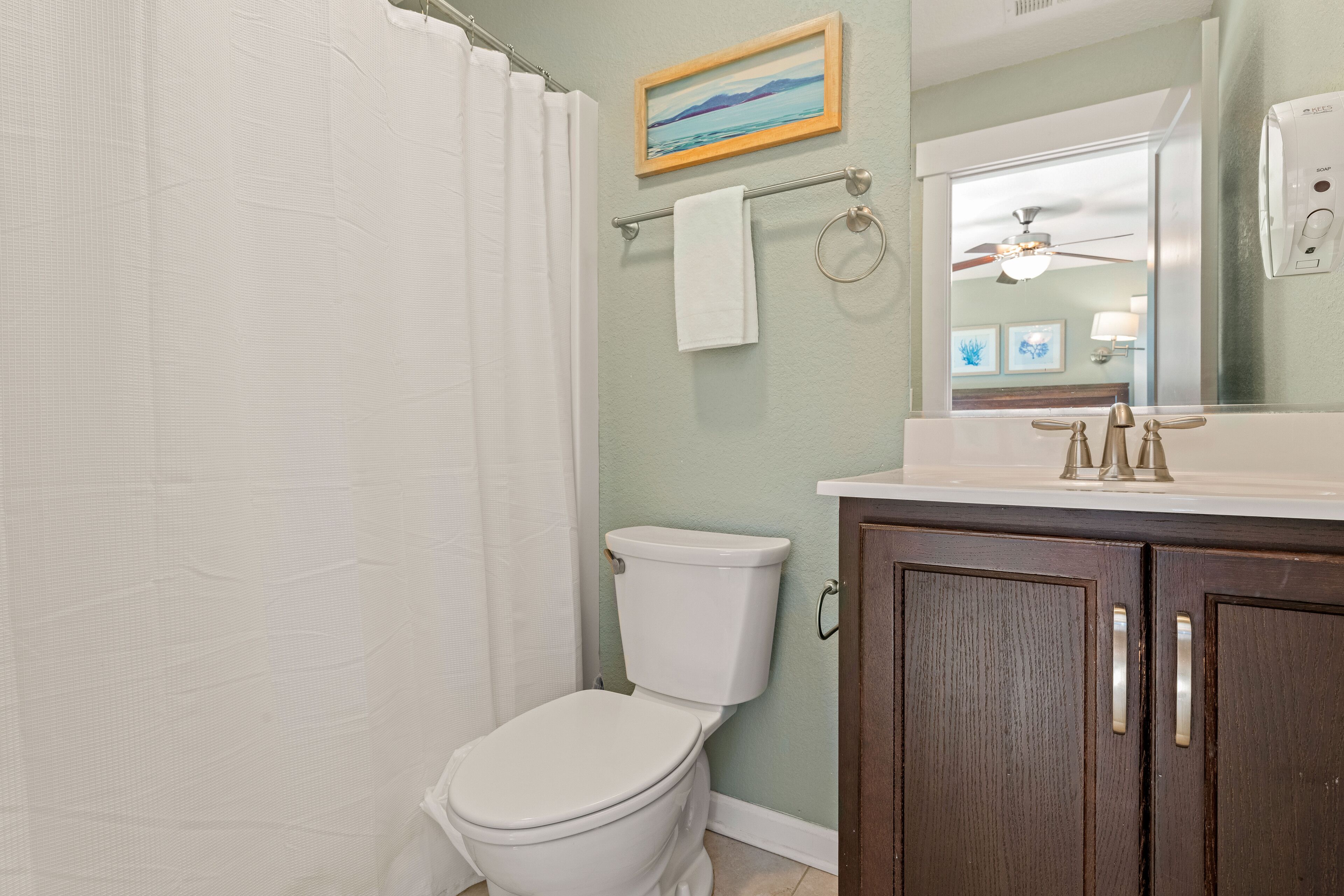 Une salle de bain équipée de toilettes, d’un rideau de douche, d’un porte-serviettes, d’un miroir et d’un ventilateur de plafond.