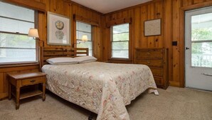 A bedroom with wooden paneled walls, a bed with a floral bedspread, two bedside lamps, a dresser, and a door with a view.