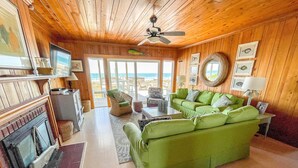 A cozy living room with a green sofa, a fireplace, and a view of the ocean. - Barnacle: Pet-Friendly Ocean Escape (Topsail Beach)