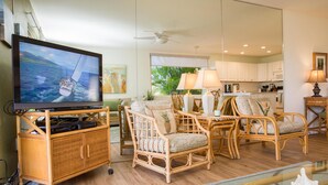 A living room with wicker furniture, a television on a stand, and a view of the kitchen and outdoors. - Hale Kamaole 177 Ground floor condo, easy access to best beach on Maui! (Kihei)