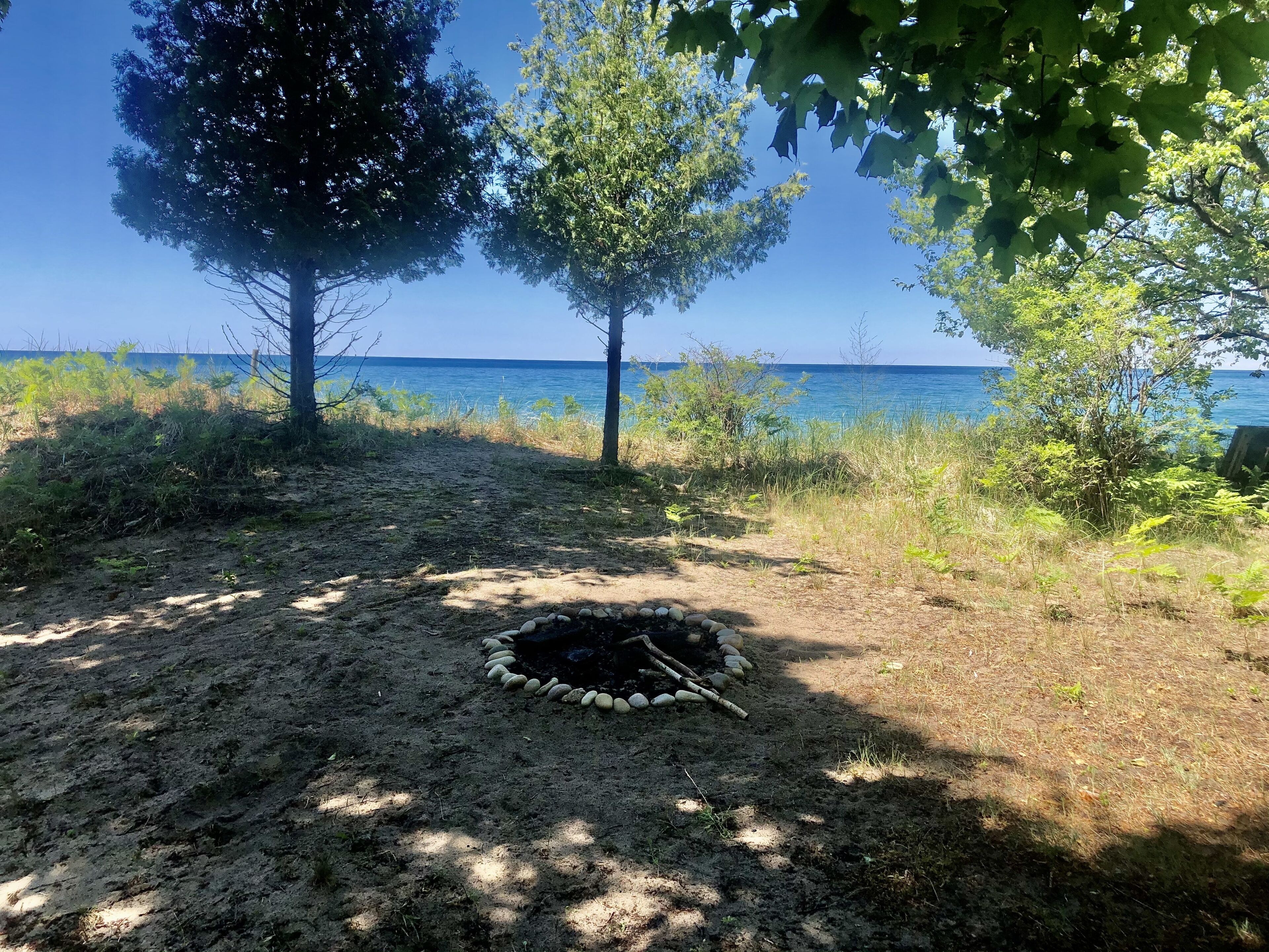 A beach scene with a fire pit surrounded by rocks, trees, and a view of the sea.