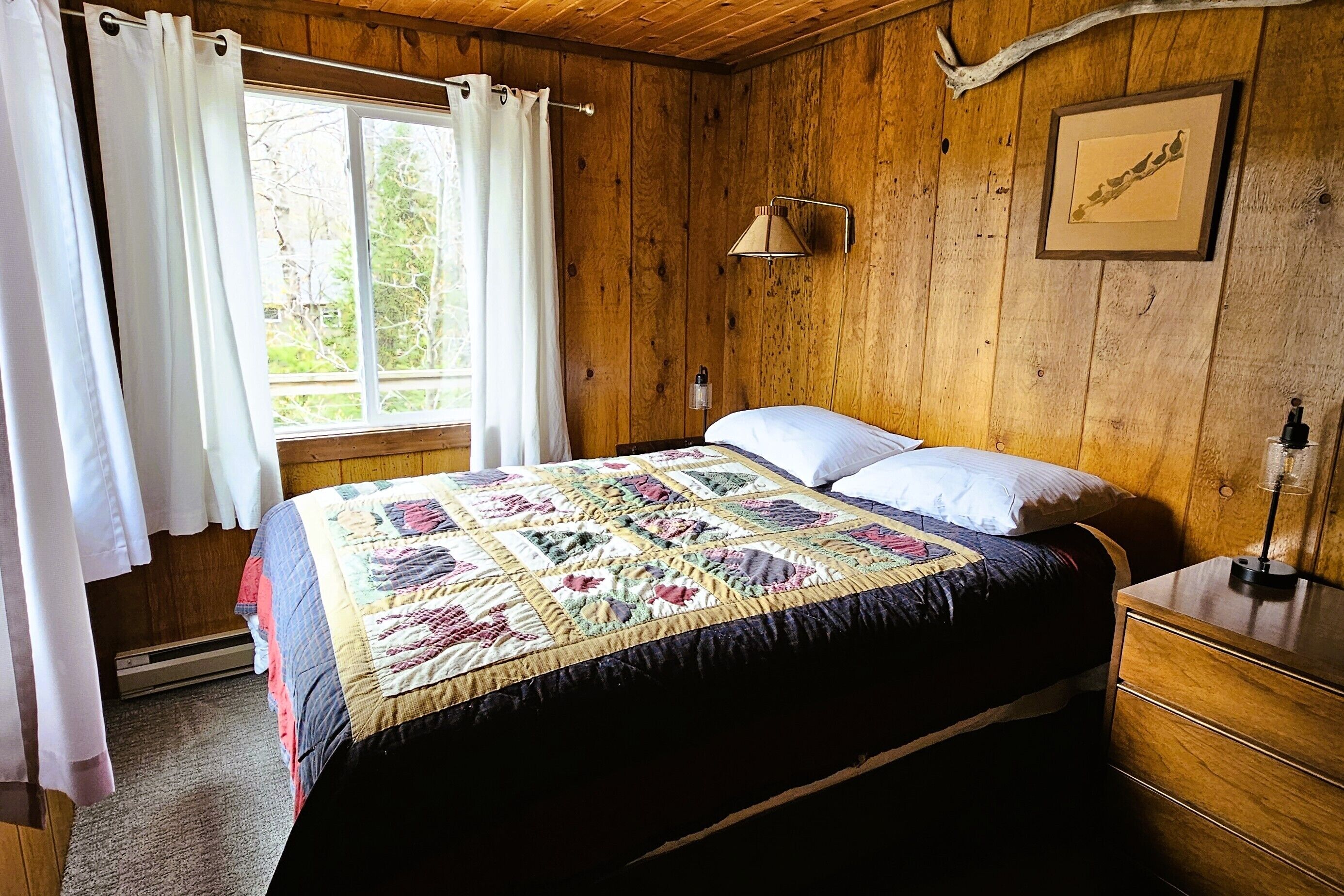 Une pièce en bois avec un lit, une table de chevet, une lampe, une fenêtre avec des rideaux et un tableau encadré au mur.