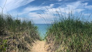 Un sentier de sable qui longe la plage et mène à la mer, bordé d’herbes hautes et sous un ciel partiellement nuageux.
