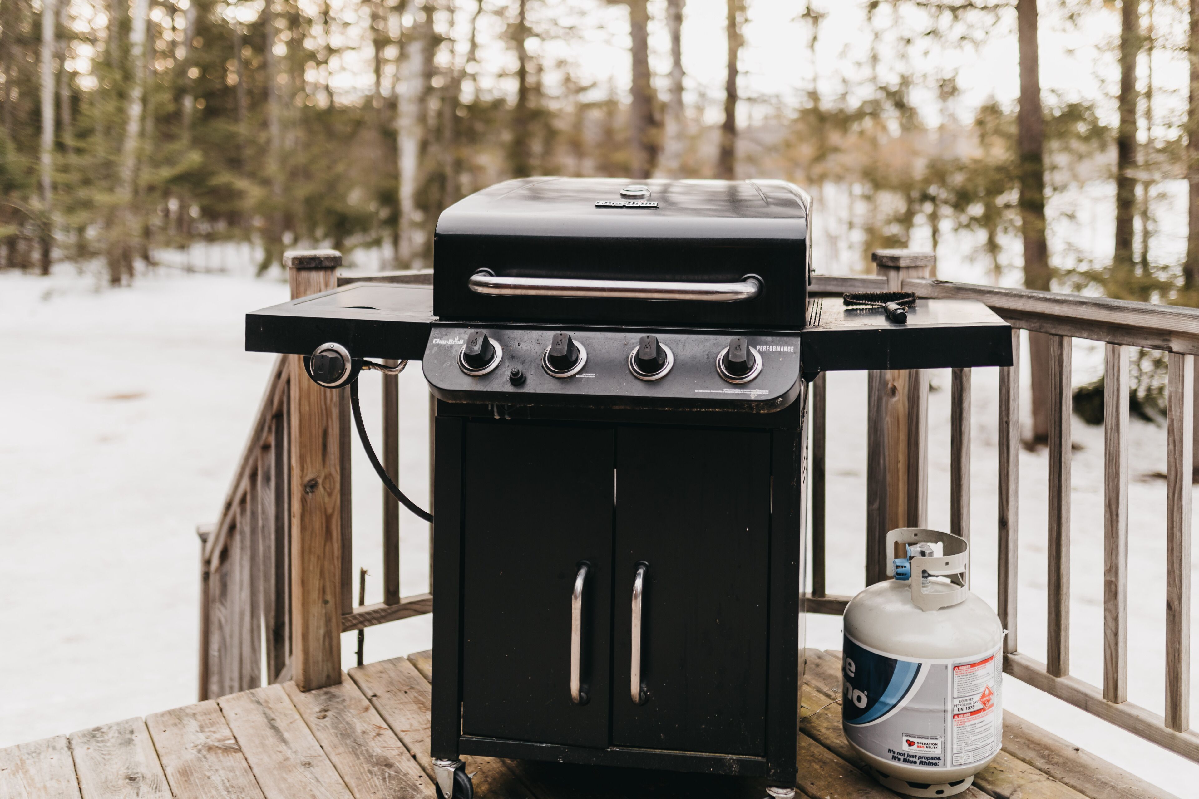 A portable charcoal grill with four burners and a gas tank on a wooden deck.