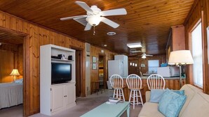 A cozy cabin interior with a wooden ceiling, a television on a white cabinet, a sofa with a cushion, and a dining area with chairs and a table. - Historic Cottage Across the Street from Beach (Carolina Beach)
