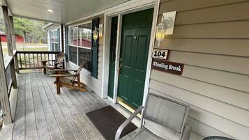 A porch with wooden chairs and a bench, a green door, and a house number sign.