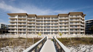 Un hotel de varios pisos con balcones, una playa de arena frente a él y un camino de madera que lleva a la entrada.
